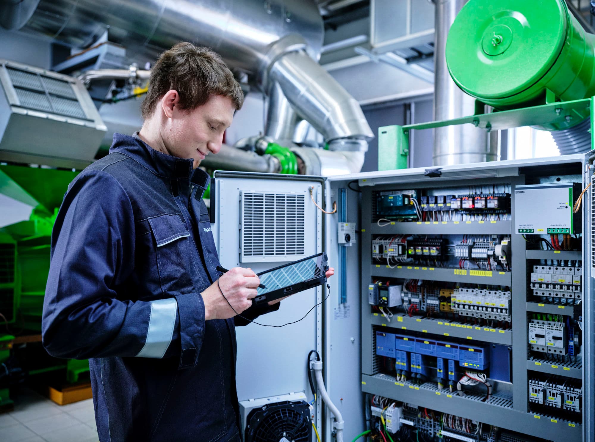 A technician in a dark uniform using a tablet to inspect or configure an open industrial control cabinet, surrounded by machinery and metal pipes.