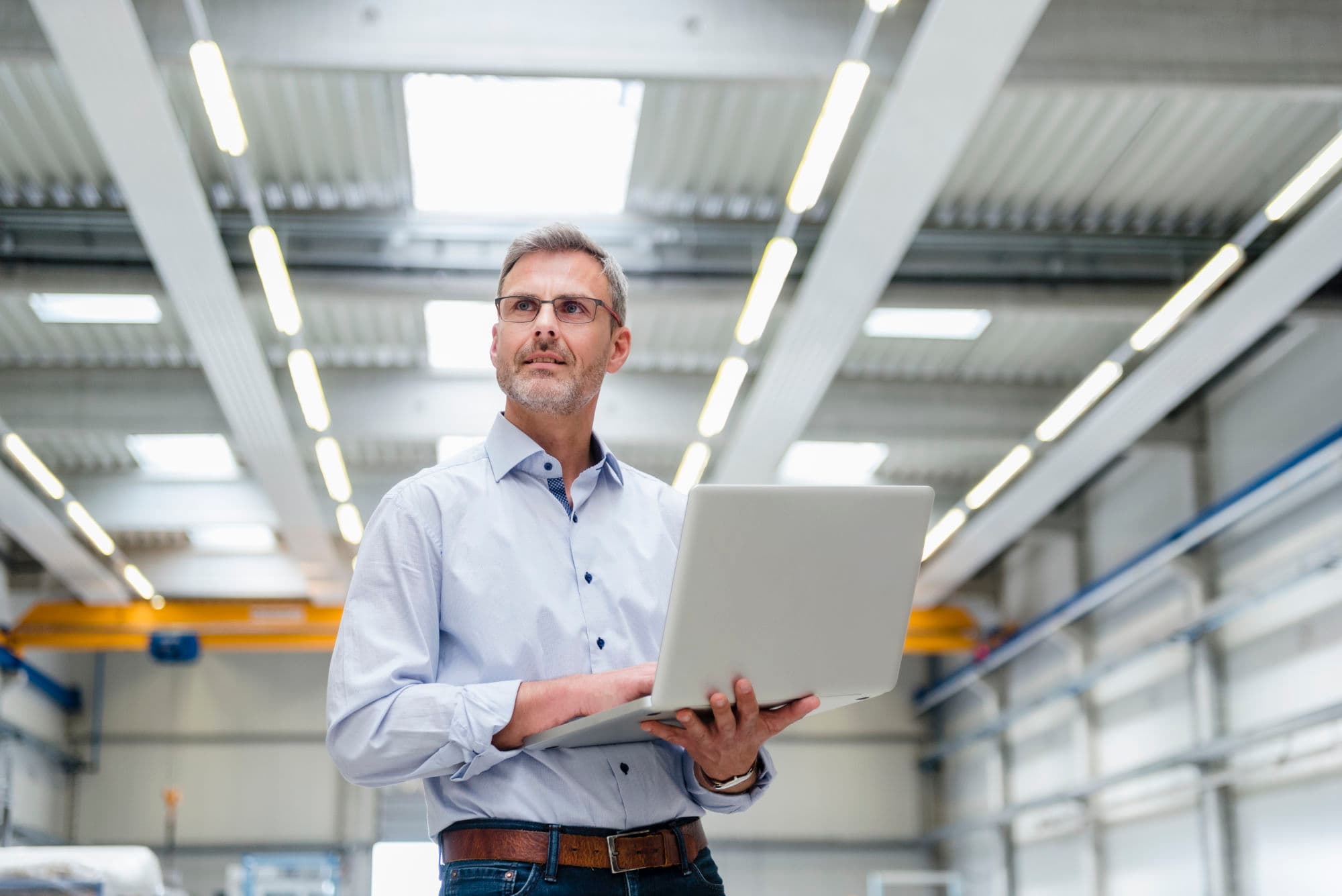 A man in a light blue shirt holding a laptop, standing in a modern industrial facility with high ceilings and bright lighting.