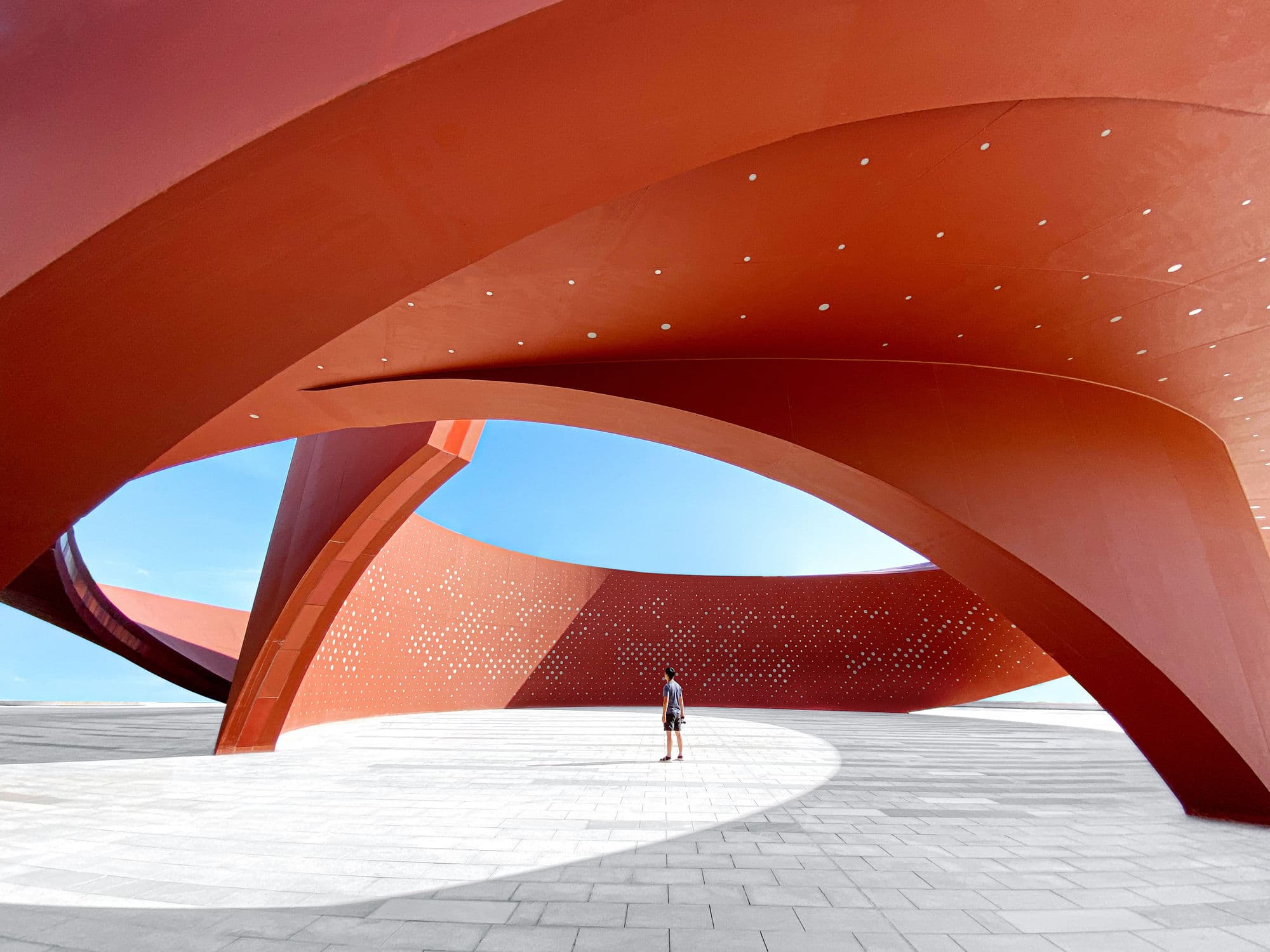 A person stands alone in a spacious architectural structure with sweeping, curved red beams and perforated walls.