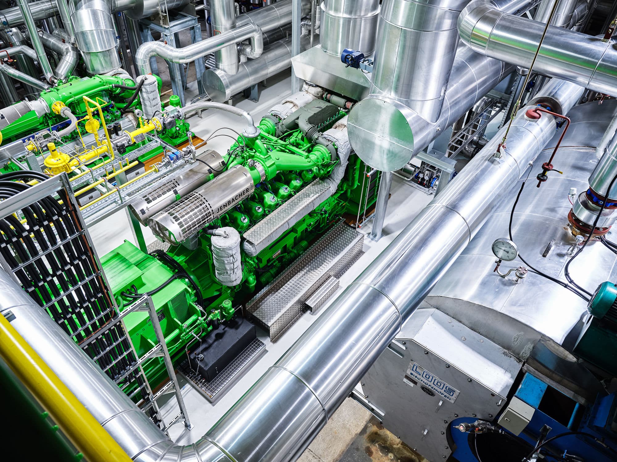 A high-tech industrial facility featuring a green INNIO Jenbacher gas engine surrounded by large silver pipelines, valves, and gauges in a clean, well-lit environment