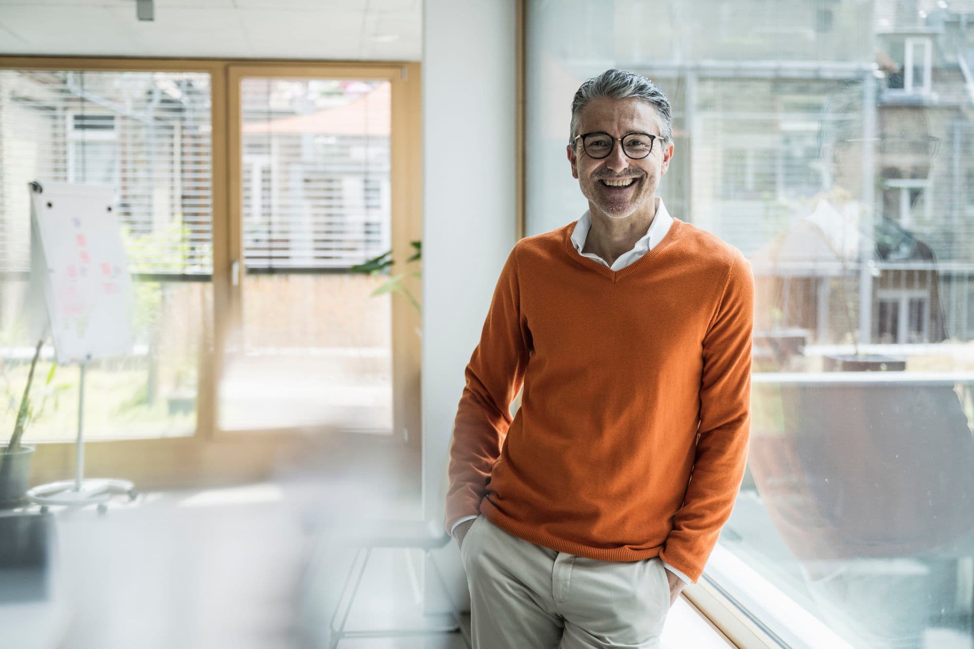 A smiling man in glasses and an orange sweater leans casually against a window in a bright, modern office with a flip chart and large windows in the background.