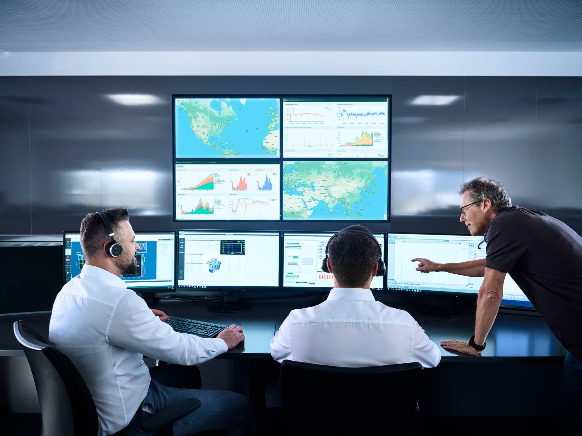Three men working in a control room with multiple computer monitors displaying data charts, maps, and system dashboards; one is standing and pointing at a screen while the others wear headsets.