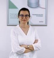 Hande Sevinç standing with arms crossed, wearing glasses and a white blouse, in front of a framed poster